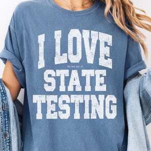 Könnte beinhalten: Blaues Jeans-T-Shirt mit dem Text "I LOVE STATE TESTING" in weißen Blockbuchstaben. Der Satz "the last day of" steht über "STATE TESTING". Das Shirt hat hochgekrempelte Ärmel und gehört zur Comfort Colors-Kollektion.