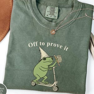 May include: A moss green crew neck t-shirt featuring a graphic of a frog wearing a pointed hat, riding a scooter, and holding a pencil and calculator. The text "Off to prove it" is printed above the frog. The shirt has a Comfort Colors label.