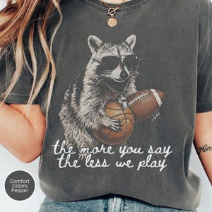 May include: A charcoal gray t-shirt featuring a graphic of a raccoon wearing sunglasses and a whistle around its neck, holding a basketball and a football. Below the graphic, the text reads, the more you say the less we play in white script.
