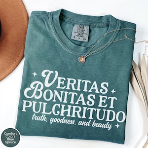 Pode incluir: Camiseta Comfort Colors azul Spruce com as palavras "Veritas Bonitas Et Pulchritudo" em branco, que significa "verdade, bondade e beleza". Um colar de ouro com um pingente repousa sobre a camiseta. Tamanho S-P-CH.