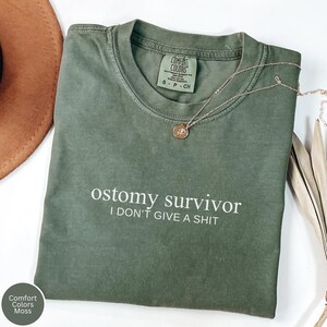 May include: Olive green Comfort Colors t-shirt with the text "ostomy survivor I DON'T GIVE A SHIT" in white. A gold necklace with a pendant is draped across the shirt. The shirt is labeled "S-P-CH".