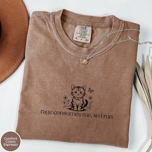 May include: Espresso-colored t-shirt with a cat graphic and the phrase "rage consumes me, so I run." The shirt features a crew neck and short sleeves. A silver necklace with a pendant is draped across the shirt.