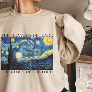 Puede incluir: Sudadera de color arena con una impresión vibrante de la pintura "La noche estrellada" de Van Gogh. La imagen incluye el texto "THE HEAVENS DECLARE THE GLORY OF THE LORD" en fuente negra. La sudadera está hecha de un material suave.