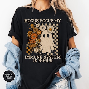 May include: Black t-shirt with the text "Hocus Pocus My Immune System Is Bogus." The shirt features a ghost graphic with flowers and a checkered pattern. The shirt is black and made of Comfort Colors material.