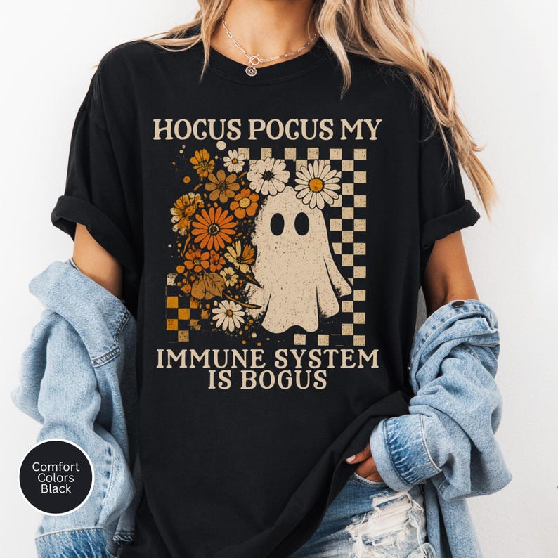 May include: Black t-shirt with the text "Hocus Pocus My Immune System Is Bogus." The shirt features a ghost graphic with flowers and a checkered pattern. The shirt is black and made of Comfort Colors material.
