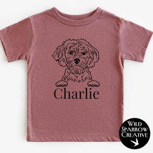 May include: A dusty rose t-shirt with a black illustration of a dog and the name "Charlie" printed below. The dog is drawn in a fluffy style. A circular logo with the text "Wild Sparrow Creative" is in the bottom right corner.