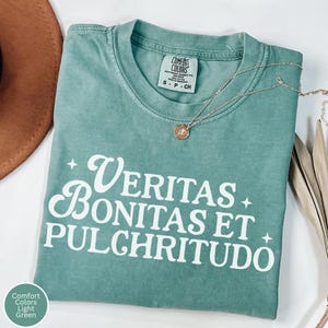 May include: Light green Comfort Colors t-shirt with the Latin phrase "Veritas Bonitas Et Pulchritudo" in white script. A gold necklace with a pendant rests on the shirt. The shirt is folded, with a brown hat visible.