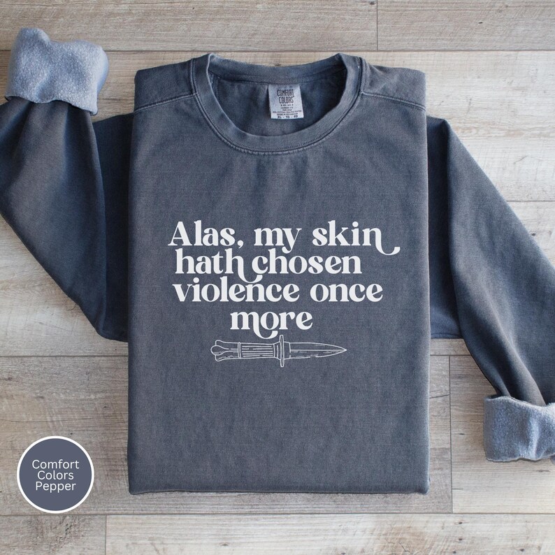 May include: A dark gray sweatshirt with the text "Alas, my skin hath chosen violence once more" and a knife graphic in white. The sweatshirt has a crew neck and long sleeves with rolled cuffs. The Comfort Colors label is visible.