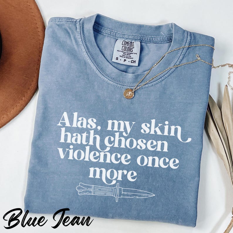 May include: A blue jean Comfort Colors t-shirt with the text "Alas, my skin hath chosen violence once more" and a knife graphic. A gold necklace with a pendant is draped on the shirt.