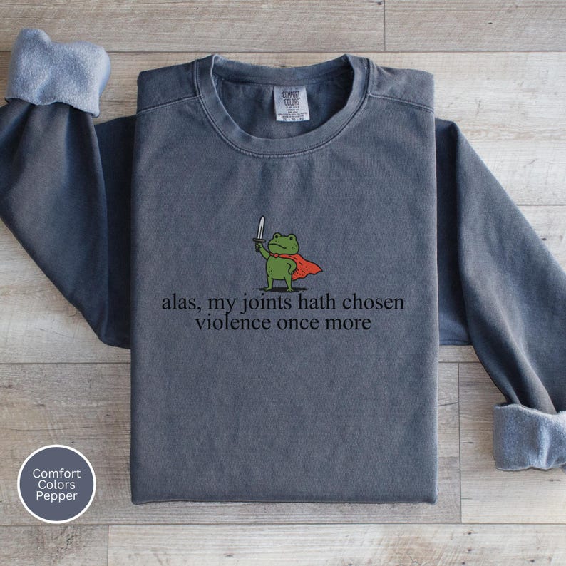 May include: A dark grey sweatshirt featuring the phrase "alas, my joints hath chosen violence once more." The design includes a cartoon frog in a red cape holding a sword. The sweatshirt is a Comfort Colors Pepper brand.