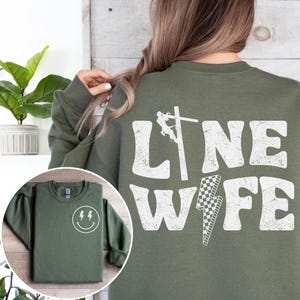 May include: Green sweatshirt with a white graphic that says "Line Wife" with a silhouette of a lineman on a power pole. The sweatshirt also has a small smiley face with lightning bolts for eyes on the left side of the chest.
