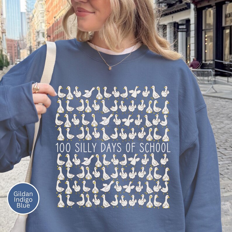 K&ouml;nnte beinhalten: Indigoblaues Sweatshirt mit einem Muster aus wei&szlig;en G&auml;nsen und dem Text "100 SILLY DAYS OF SCHOOL". Das Sweatshirt besteht aus einem weichen Material, ideal f&uuml;r die Schule.