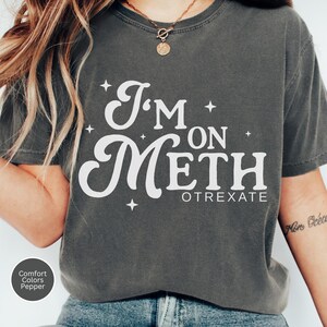 May include: Dark grey t-shirt with the text "I'm on Meth Otrexate" in white, with star accents. The shirt is a Comfort Colors Pepper tee. The shirt is a casual wear item.