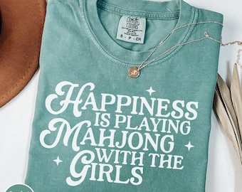 May include: A light green Comfort Colors t-shirt with the text "Happiness is playing Mahjong with the Girls" in white. A gold necklace with a pendant is on the shirt. The shirt is folded.