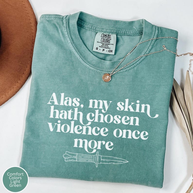 May include: Light green Comfort Colors t-shirt with the text "Alas, my skin hath chosen violence once more" and a knife graphic. A gold necklace with a pendant is draped across the shirt.
