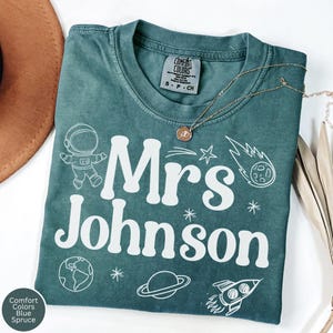 Puede incluir: Una camiseta verde azulado con las palabras "Mrs Johnson" en blanco, junto con gráficos temáticos del espacio como un astronauta, un cohete y planetas. La camiseta tiene cuello redondo y una pequeña etiqueta en la parte superior. Un collar dorado está sobre la camiseta.