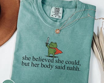 May include: Light green Comfort Colors t-shirt with a cartoon frog holding a sword and wearing a red cape. The text "she believed she could, but her body said nahh" is printed below the frog. A gold necklace is draped across the shirt.