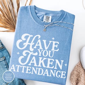 K&ouml;nnte beinhalten: Hellblaues Comfort Colors Washed Denim T-Shirt mit dem wei&szlig;en Schriftzug &bdquo;Have You Taken Attendance&ldquo;. Eine silberne Halskette mit Sonnenblumenanh&auml;nger liegt auf dem Shirt. Das Shirt ist auf einer wei&szlig;en Oberfl&auml;che gefaltet.
