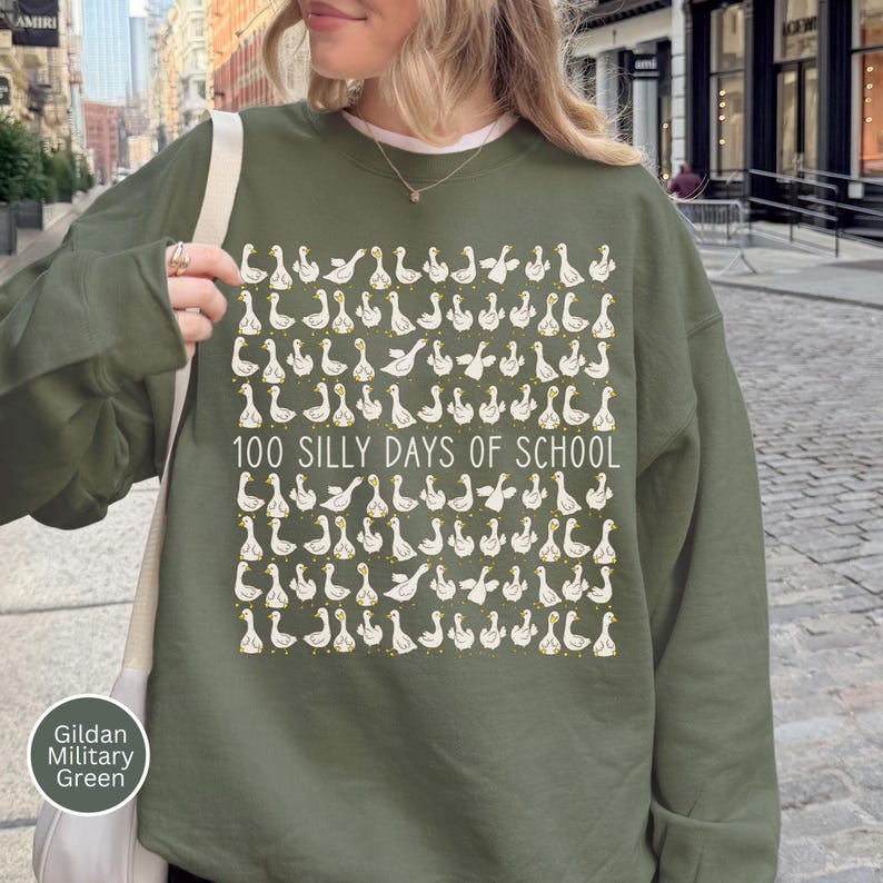 K&ouml;nnte beinhalten: Olivgr&uuml;nes Sweatshirt mit einem sich wiederholenden Muster aus wei&szlig;en G&auml;nsen und dem Text "100 SILLY DAYS OF SCHOOL" in Wei&szlig;. Das Sweatshirt hat die Farbe Gildan Military Green.