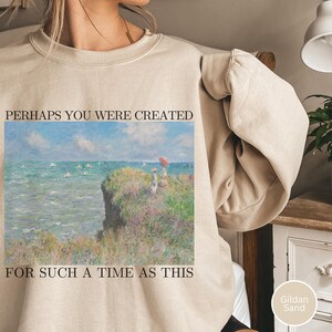 Puede incluir: Sudadera color arena con una pintura de un paisaje costero y el texto "PERHAPS YOU WERE CREATED FOR SUCH A TIME AS THIS." La obra de arte representa un acantilado con figuras, el océano y un cielo azul. La sudadera es de color Gildan Arena.