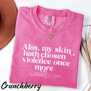 May include: A pink t-shirt with the text "Alas, my skin hath chosen violence once more" and a knife graphic. The shirt has a crew neck and a small tag. A gold necklace with a pendant is draped across the shirt. The word "Crunchberry" is at the bottom.
