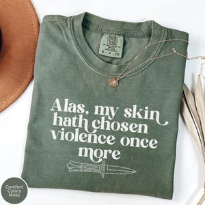 May include: A moss green Comfort Colors t-shirt with the text "Alas, my skin hath chosen violence once more" and a knife graphic. A gold necklace with a pendant is draped across the shirt. A brown hat is visible in the background.