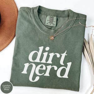 May include: A moss green short-sleeve t-shirt with the text "dirt nerd" printed in white on the front. The shirt features a Comfort Colors brand label at the inner neckline, indicating a size small, and is paired with a thin gold-tone necklace.