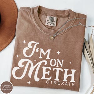 May include: Espresso-colored Comfort Colors t-shirt with the text "I'm on Meth" in a white, retro font. The shirt has a crew neck and short sleeves. A silver necklace with a small pendant is draped across the shirt.