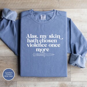 May include: Blue jean-colored sweatshirt with the text "Alas, my skin hath chosen violence once more" and a knife graphic. The long-sleeved shirt has a crew neck and rolled cuffs. The Comfort Colors brand is visible.