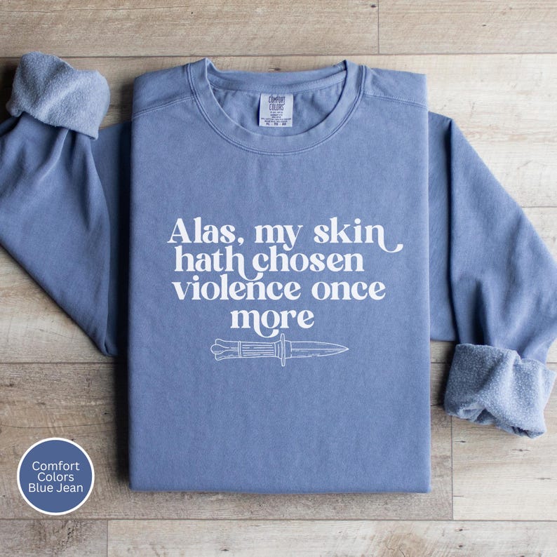 May include: Blue jean-colored sweatshirt with the text "Alas, my skin hath chosen violence once more" and a knife graphic. The long-sleeved shirt has a crew neck and rolled cuffs. The Comfort Colors brand is visible.