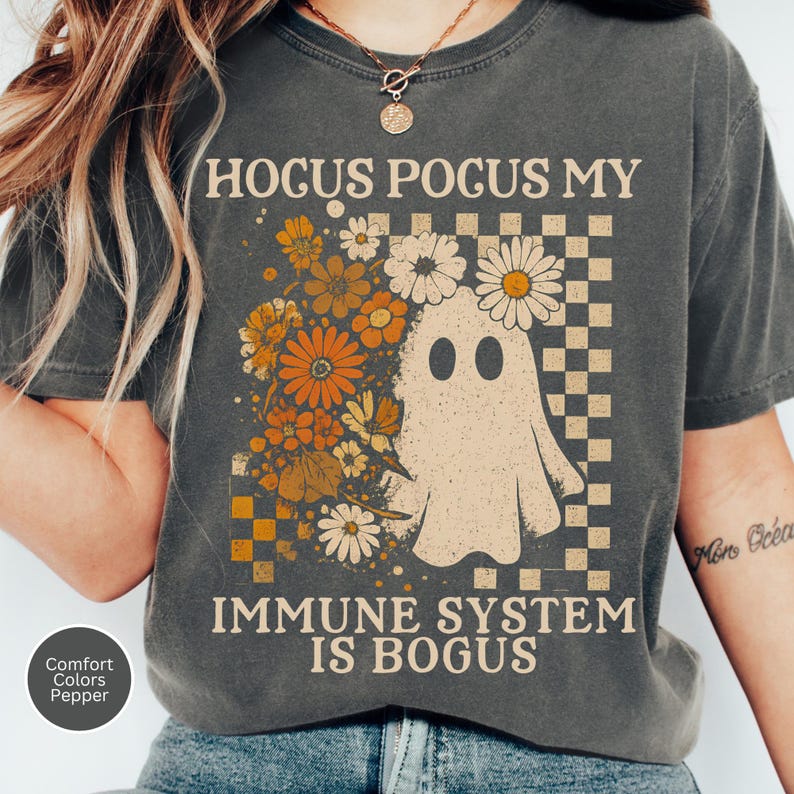 May include: Dark grey t-shirt with the text "Hocus Pocus My Immune System Is Bogus." The shirt features a ghost graphic with flowers and a checkered pattern. The shirt is a Comfort Colors Pepper tee.
