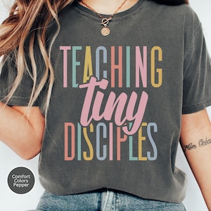 May include: Dark gray t-shirt with the words "TEACHING tiny DISCIPLES" in colorful block letters. The shirt is a Comfort Colors Pepper tee.