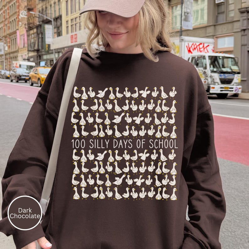 K&ouml;nnte beinhalten: Dunkel schokoladenbraunes Sweatshirt mit einem Muster aus wei&szlig;en und gelben Cartoon-G&auml;nsen. Der Text "100 SILLY DAYS OF SCHOOL" ist darunter gedruckt. Das Sweatshirt ist ein l&auml;ssiger Stil.