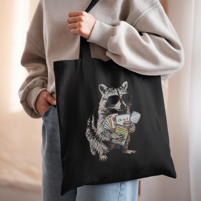 May include: A black tote bag featuring a graphic of a raccoon wearing sunglasses and holding a set of educational flashcards. The cards display the text ch-a-t, Phonics Fun & Speech Sounds, and various letters, along with a small handheld mirror.