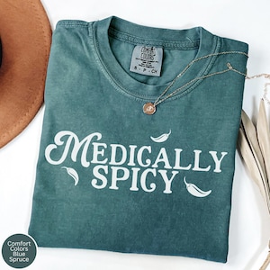 May include: A blue spruce Comfort Colors t-shirt with the words "Medically Spicy" in white script, accompanied by chili pepper graphics. A gold necklace with a pendant is draped across the shirt.