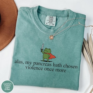 May include: A light green short-sleeve t-shirt featuring a graphic of a green frog wearing a red cape and holding a sword. The text reads, "alas, my pancreas hath chosen violence once more." The shirt has a Comfort Colors brand label at the neck.