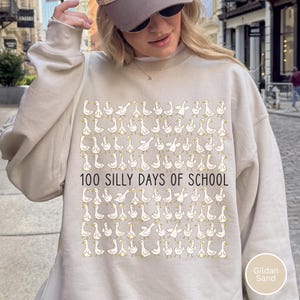K&ouml;nnte beinhalten: Beiges Sweatshirt mit einem sich wiederholenden Muster aus wei&szlig;en Cartoon-G&auml;nsen und dem Text "100 SILLY DAYS OF SCHOOL" in Schwarz. Das Sweatshirt hat die Farbe Gildan Sand.