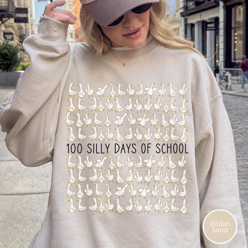 K&ouml;nnte beinhalten: Beiges Sweatshirt mit einem sich wiederholenden Muster aus wei&szlig;en Cartoon-G&auml;nsen und dem Text "100 SILLY DAYS OF SCHOOL" in Schwarz. Das Sweatshirt hat die Farbe Gildan Sand.