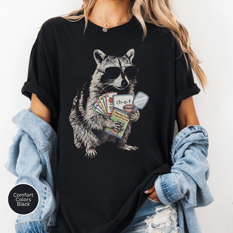 May include: A black t-shirt featuring a graphic of a raccoon wearing sunglasses and holding a set of educational flashcards. The cards display the letters a, b, c, and the text ch-a-t with an illustration of a mouth and a handheld mirror.