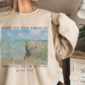 Puede incluir: Sudadera color arena con una pintura de paisaje costero. La obra de arte muestra un acantilado con figuras, el océano y un cielo azul. El texto dice: "WHEN YOU PASS THROUGH THE WATERS I WILL BE WITH YOU Isaiah 43:2".