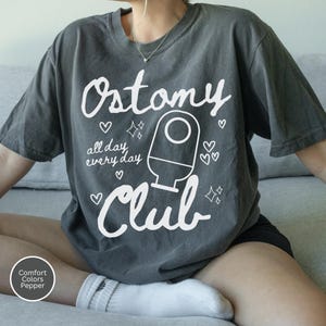 May include: Dark grey t-shirt with the words "Ostomy Club" in white script, along with an illustration of a medical device and the phrase "all day every day." The design includes hearts and stars. The shirt is a Comfort Colors Pepper tee.