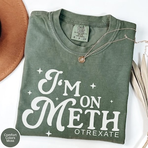 May include: A moss green Comfort Colors t-shirt with the text "I'm on Meth Otrexate" in white. A gold necklace with a pendant is draped across the shirt. A brown hat is visible in the upper left corner.