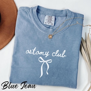 May include: A blue jean colored t-shirt with the words "ostomy club" in white script and a white bow graphic. A gold necklace with a pendant is draped across the shirt. The shirt has a Comfort Colors tag.