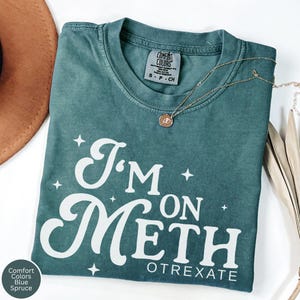May include: Blue spruce Comfort Colors t-shirt with the text "I'm on Meth" in white script. A gold necklace with a pendant is draped across the shirt. The shirt is folded on a white surface.