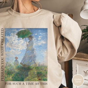 Puede incluir: Sudadera color arena con una imagen impresa de una mujer con un paraguas verde en un campo, que recuerda a una pintura clásica. El texto "PERHAPS YOU WERE CREATED FOR SUCH A TIME AS THIS" está impreso en el lado izquierdo.