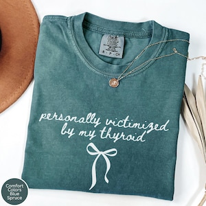 May include: Blue spruce Comfort Colors t-shirt with the text "personally victimized by my thyroid" in white script. A white bow graphic is below the text. A gold necklace with a pendant is on the shirt.