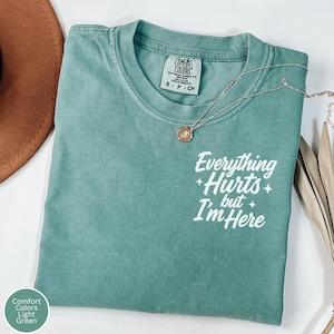 Pode incluir: Camiseta Comfort Colors verde claro com o texto branco "Everything Hurts but I'm Here". Um colar de ouro com um pingente está sobre a camiseta. A camiseta está dobrada e tem uma etiqueta que diz "S-P-CH".