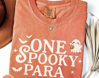 May include: A coral-colored t-shirt with the text "ONE SPOOKY PARA" in white, along with bat and star graphics. A cute ghost holding a pumpkin is also featured. A gold necklace is draped across the shirt.