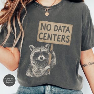 以下が含まれることがあります： サングラスをかけ、NO DATA CENTERSと黒い大文字で書かれた段ボールの看板を持つアライグマのグラフィックがプリントされたチャコールグレーのTシャツ。クルーネックで半袖、ヴィンテージ風のウォッシュ加工が施されています。