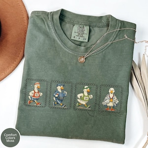 May include: A moss green Comfort Colors t-shirt featuring a series of four cartoon ducks in running attire, each within a postage stamp frame. The shirt includes a small tag at the neckline and a silver necklace.